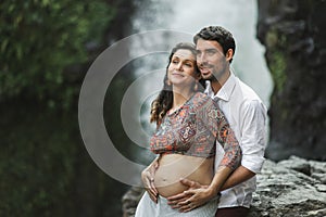 Young pregnant happy couple with view of waterfall. Pregnancy travel lifestyle.