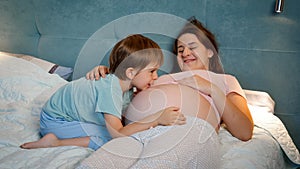 Young pregnant mother with big belly lying with hr older son in bed at night. Family having good time together and