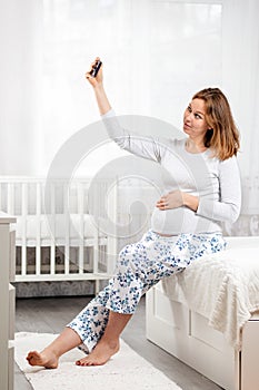 A young pregnant smiling woman is sitting on a bed and taking a selfie on her smartphone. Concept of social networks and pregnancy