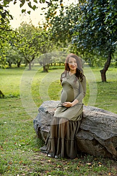 Young pregnant woman in anticipation of a baby sitting on a big stone in a park in the sun rays