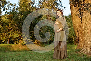 Young pregnant woman in anticipation of a baby standing in a park in the sun rays