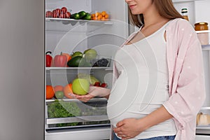 Young pregnant woman with apple near fridge at home, closeup. Healthy eating