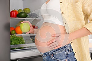 Young pregnant woman with apple near fridge at home, closeup. Healthy eating