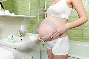Young pregnant woman applying anti-stretch marks cream to the abdomen in the morning in the bathroom. Pregnancy, maternity and