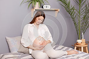 Young pregnant woman with bare belly sitting in bedroom, enjoying peaceful moment while awaiting baby's arrival