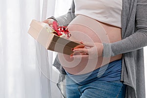 Young pregnant woman with big belly holding a gift wrapped with red ribbon, baby shower party with presents.