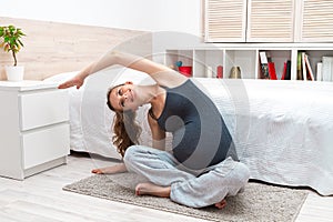 Young pregnant woman with big belly sitting on mat cross-legged and stretching yoga exercises.