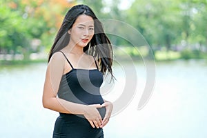 Young pregnant woman with black dress stand and touch her belly stand in front of lake with day light