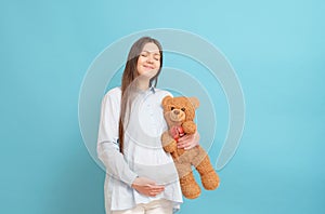 young pregnant woman on blue background in blue clothes with teddy bear in hands
