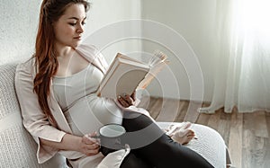 Young pregnant woman with a book and a cup is resting on the couch at home