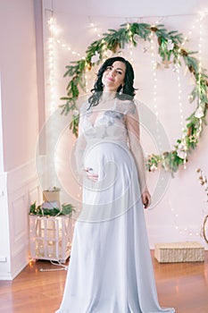 Young pregnant woman with dark hair in airy dress in a room decorated with pine needles and sparkling garlands for Christmas