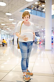 Young pregnant woman in despair because of the untied laceup in the hall of the shopping center. Problems of pregnant women with photo