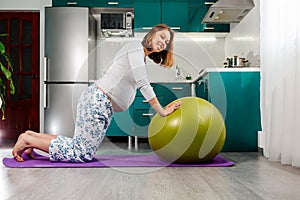 A young pregnant woman does an exercise with a fitball on a rug in the kitchen. Concept of sports and health during pregnancy