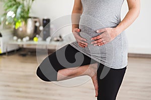 Young pregnant woman standing in tree pose doing prenatal yoga holding belly