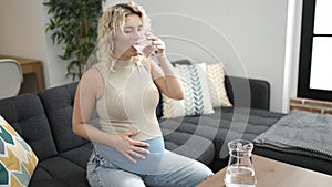 Young pregnant woman drinking glass of water sitting on sofa at home