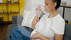 Young pregnant woman drinking glass of water sitting on sofa at home