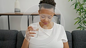 Young pregnant woman drinking glass of water sitting on sofa at home