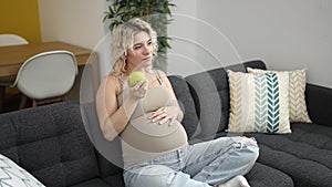 Young pregnant woman eating apple touching belly at home