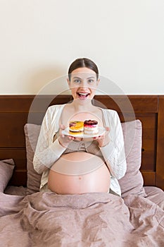Young pregnant woman enjoys eating tasty cake resting in bed at home. Unhealthy diet during pregnancy concept