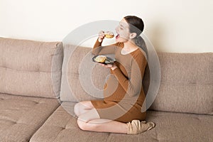Young pregnant woman enjoys eating tasty cake resting on the sofa at home. Unhealthy diet during pregnancy concept