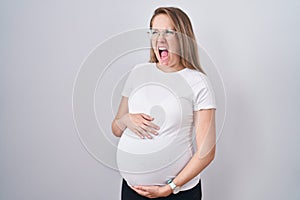 Young pregnant woman expecting a baby, touching pregnant belly angry and mad screaming frustrated and furious, shouting with anger