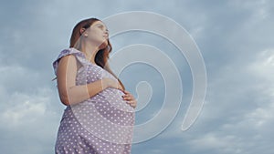 Young pregnant woman folded her hands in pray to God standing alone in field against of cloudy sky, future mother walking on natur