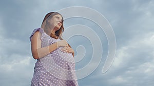 Young pregnant woman folded her hands in pray to God standing alone in field against of cloudy sky, future mother walking on natur