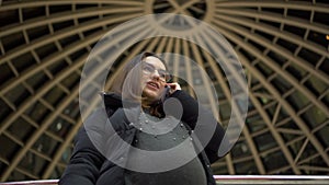 A young pregnant woman in glasses and a jacket is standing and talking on the phone. A girl in a shopping center uses a