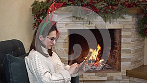 A young pregnant woman with glasses sits in a chair by the fireplace with horns on her head and writes on the tablet.