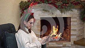 A young pregnant woman with glasses sits in a chair by the fireplace with horns on her head and writes on the tablet.