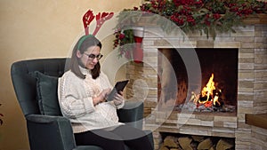 A young pregnant woman with glasses sits in a chair by the fireplace with horns on her head and writes on the tablet.