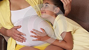 Young pregnant woman and her daughter are hugging while sitting on the couch. The girl puts her ear to the belly of a