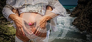 Young pregnant woman with her hands and those of her partner making a heart in her belly on the beach