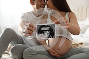 Young pregnant woman with her husband in bedroom, focus on ultrasound picture of baby
