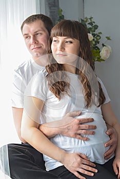 Young pregnant woman and her husband are hugging, touching the belly near window and looking ouside