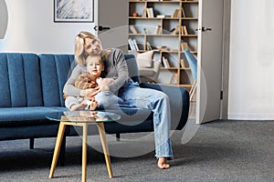 Young pregnant woman and her son sit on sofa at home, little boy spend time together, enjoy communication feeling kicks of unborn