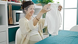 Young pregnant woman holding baby clothes sitting on table at dinning room