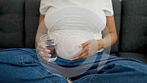 Young pregnant woman holding glass of water sitting on sofa at home