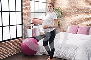 Young pregnant woman holding pilate mat at bedroom afraid and shocked with surprise and amazed expression, fear and excited face