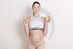 Young pregnant woman holding up two halves of fresh avocado while posing against white wall, expectant mother showing her bare