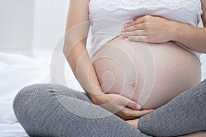 A young pregnant woman holds her hands on her swollen belly. Happy pregnancy sitting on the bed at home in the bedroom. pregnant