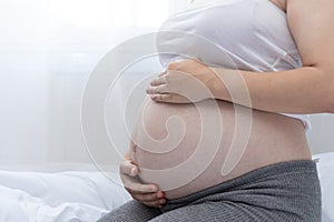 A young pregnant woman holds her hands on her swollen belly. Happy pregnancy sitting on the bed at home in the bedroom. pregnant