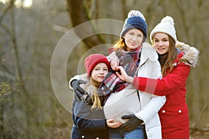Young pregnant woman hugging her older daughters. Older siblings having fun with her pregnant mom outdoors. Mother and her kids