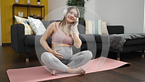 Young pregnant woman listening to music sitting on yoga mat at home