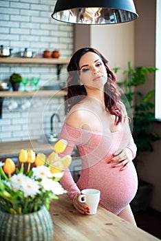 Young pregnant woman in pink bodysuit drinking water in the kitchen. How to get rid of morning sickness and toxemia. Drugs and