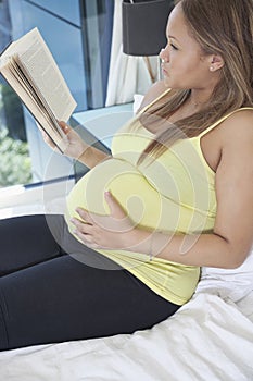 Young pregnant woman reading book while relaxing on bed at home