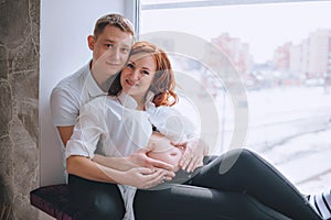 Young pregnant woman with red hair sits on the windowsill. Hugs her husband. He was hugging a naked pregnant belly.