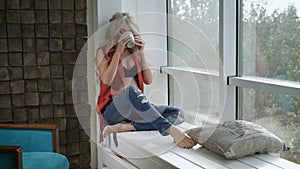 Young pregnant woman in red shirt sitting on the windowsill with the cup of tea. Beautiful future mother drinking tea