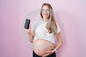 Young pregnant woman showing belly holding smartphone smiling looking to the side and staring away thinking