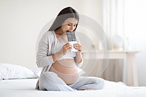 Young pregnant woman sitting on bed, eating chocolate